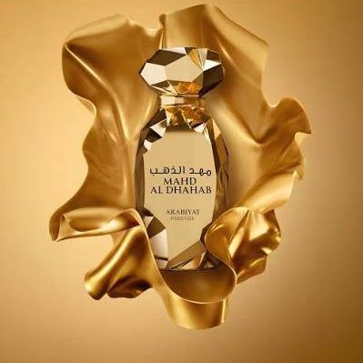 A perfume bottle with a faceted crystal-like cap and label, wrapped in flowing metallic gold fabric against a warm gold background. The label reads 'MAHDI AL DHHABAB' and 'ARABIYAT'.